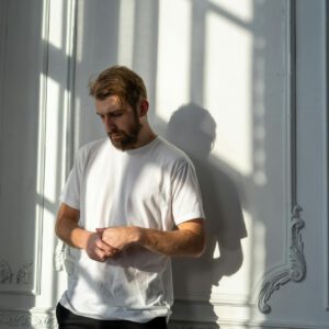 A thoughtful bearded man in a white t-shirt stands in a sunlit room with ornate walls.